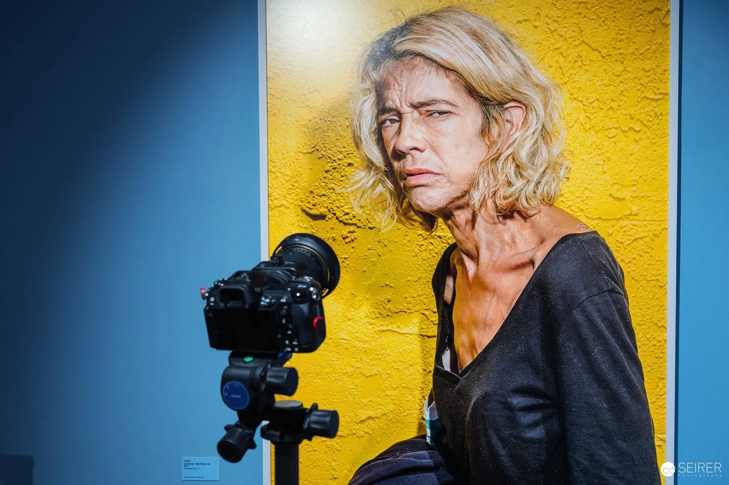 Ein Porträtfoto eines älteren Mannes mit lockigem, blondem Haar vor einer gelben Wand, aufgenommen durch eine Kamera.